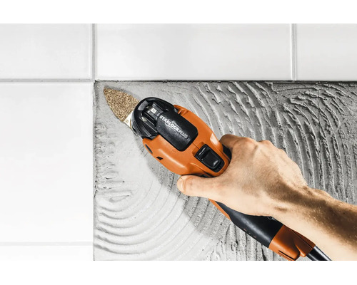 Ein Multifunktionswerkzeug mit Starlock Plus Aufnahme schleift Fliesenkleber neben einer Wand aus weißen Fliesen.