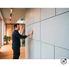 Ein Mann berührt eine Wandverkleidung aus quadratischen Platten in einem modernen Wohnraum mit Holzboden und Sichtbetondecke. (Image AI modified or generated)