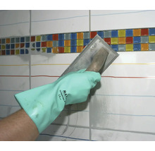 Eine Hand mit Schutzhandschuh verteilt mit einer Glättkelle Fugenmörtel auf einer Fliesenwand mit einem Mosaikbänderdekor.