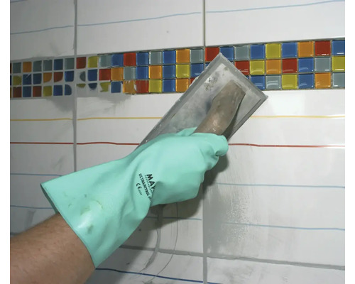 Eine Hand mit Schutzhandschuh verteilt mit einer Glättkelle Fugenmörtel auf einer Fliesenwand mit einem Mosaikbänderdekor.