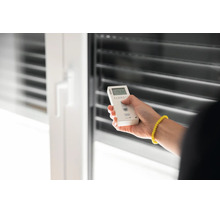 Eine Hand bedient eine Fernbedienung für Rollläden vor einem Fenster mit Jalousien.