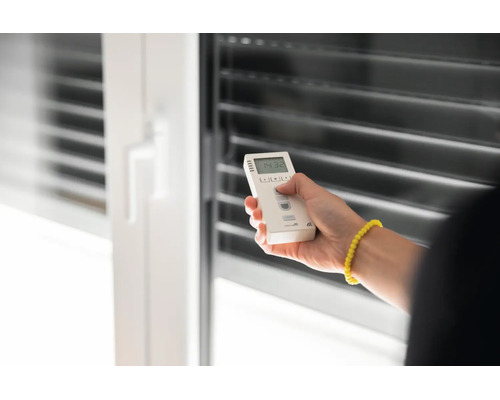 Eine Hand bedient eine Fernbedienung für Rollläden vor einem Fenster mit Jalousien.