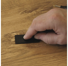 Eine Hand benutzt einen schwarzen Schaber, um Reparaturwachs auf eine Holzdielenoberfläche aufzutragen, um eine Schadstelle auszubessern.
