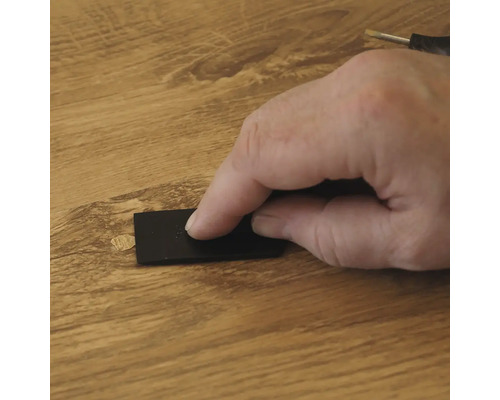 Eine Hand benutzt einen schwarzen Schaber, um Reparaturwachs auf eine Holzdielenoberfläche aufzutragen, um eine Schadstelle auszubessern.