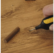 Hand der einen Wachsschmelzer über einer Holzoberfläche hält neben einem Wachsstab zur Reparatur von Bodenbelägen
