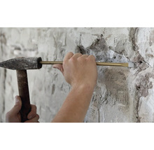 Handwerker bei der Arbeit mit einem Schlosserhammer und einem Meißel zum Abtragen von Mauerwerk an einer verputzten Wand.