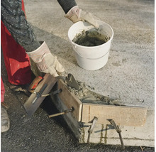 Handwerker beim Betonieren einer Mauer mit Kelle und Eimer sowie Holzschalung auf einer Baustelle.