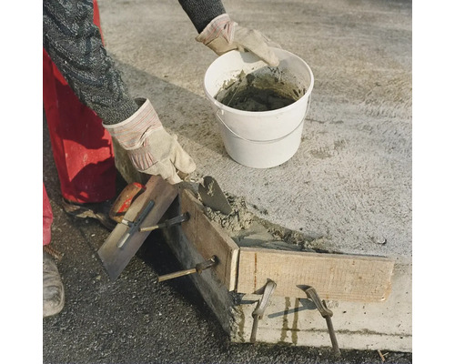 Handwerker beim Betonieren einer Mauer mit Kelle und Eimer sowie Holzschalung auf einer Baustelle.