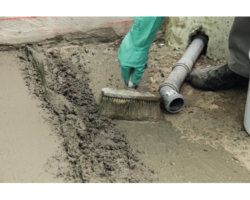 Handwerker streicht frischen Betonestrich mit einer Bürste auf einem Boden neben einem Abwasserrohr glatt.