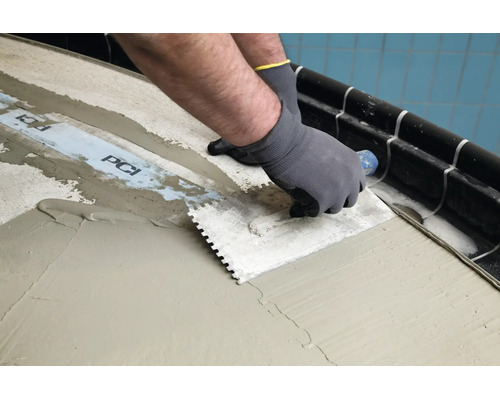 Handwerker beim Auftragen von Fliesenkleber mit einer Zahnkelle auf PCI Dichtband am Boden.