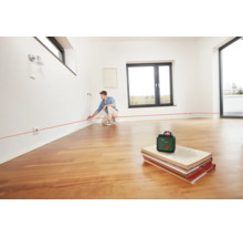 Ein Mann nutzt einen Bosch Kreuzlinienlaser in einem hellen Raum mit Parkettboden, um eine horizontale Linie an der Wand zu markieren.