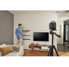 Mann richtet Wandregale in einem Wohnzimmer aus mithilfe eines auf einem Stativ montierten Bosch Kreuzlinienlasers mit roter Laserlinie.