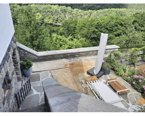 Eine Terrasse aus Naturstein mit Sitzmöbeln aus Holz, Sonnenschirm und Blick auf den Wald bei Bauarbeiten mit Sand auf dem Boden.