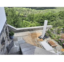 Eine Terrasse aus Naturstein mit Sitzmöbeln aus Holz, Sonnenschirm und Blick auf den Wald bei Bauarbeiten mit Sand auf dem Boden.