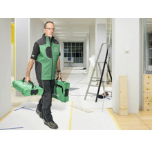 Ein Handwerker trägt zwei Werkzeugkoffer durch einen hellen, im Umbau befindlichen Raum mit einer Leiter.