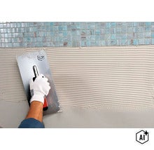 Eine Hand in einem Handschuh trägt mit einer Zahnkelle Fliesenkleber auf eine Wand unterhalb von Mosaikfliesen auf. (Image AI modified or generated)