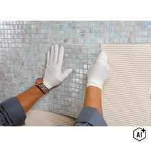 Handwerker mit Handschuhen beim Verlegen von kleinteiligen Glasmosaikfliesen auf eine mit Fliesenkleber eingestrichene Wandfläche (Image AI modified or generated)