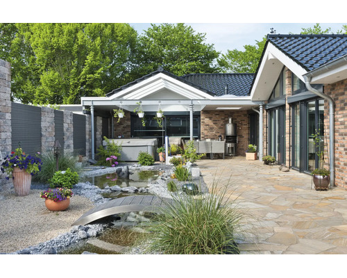 Gepflegte Terrasse mit Natursteinboden, kleinem Gartenteich mit Brücke, Whirlpool und überdachtem Sitzbereich vor einem modernen Backsteinhaus.