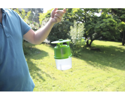 Eine Person hält eine Insektenfalle für den Außenbereich in die Höhe vor einem Garten.
