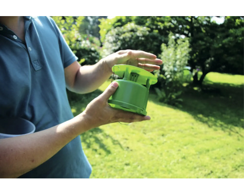 Eine Person hält eine grüne Insektenfalle in den Händen vor einem Gartenhintergrund.
