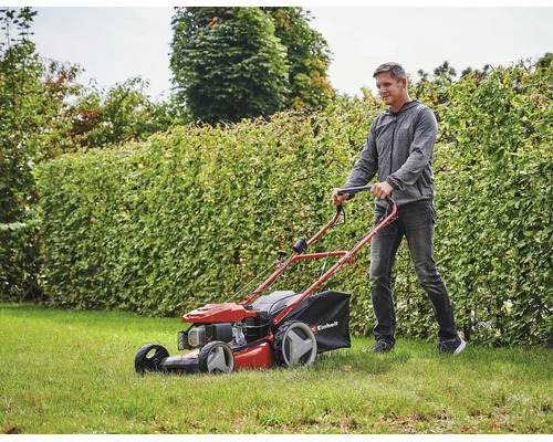 Ein Mann mäht Rasen mit einem benzinbetriebenen Rasenmäher von Einhell vor einer Hecke.