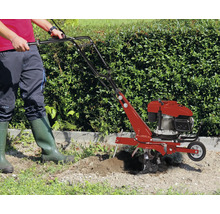 Eine Person benutzt eine benzinbetriebene Motorhacke im Garten vor einer Hecke.