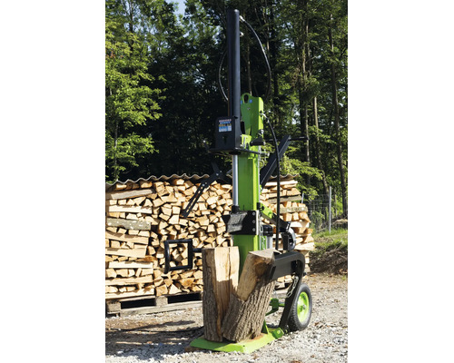 Ein vertikaler Holzspalter steht vor einem Holzstapel auf einem Waldweg.