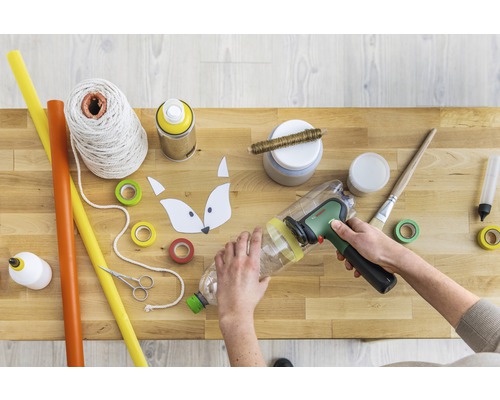 Basteltisch mit Bastelmaterialien und einer Person, die eine Plastikflasche bearbeitet.