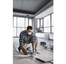 Ein Mann kniet in einem Raum und schneidet eine Fliese mit einer Schleifmaschine. Er trägt eine Schutzmaske und eine Schutzbrille.