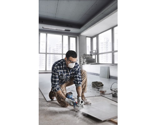 Ein Mann kniet in einem Raum und schneidet eine Fliese mit einer Schleifmaschine. Er trägt eine Schutzmaske und eine Schutzbrille.