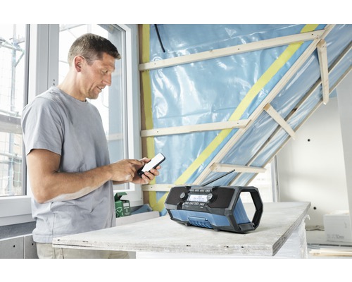 Mann bedient ein Mobiltelefon neben einem Baustellenradio in einem Raum mit unfertigen Wänden