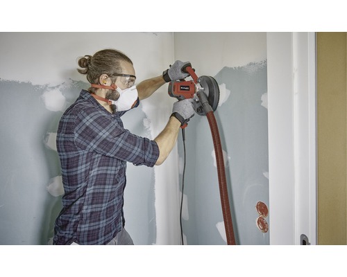 Ein Mann schleift eine Wand mit einem Trockenbauschleifer in einem Raum.