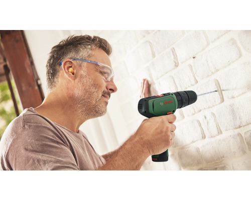 Ein Mann bohrt mit einer Bosch Bohrmaschine in eine Ziegelwand und trägt dabei Schutzbrille und Ohrenstöpsel.