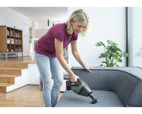 Frau saugt mit einem Akkusauger ein graues Sofa in einem hellen Wohnzimmer.