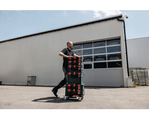 Mann transportiert Werkzeugkoffer auf Rollen vor einem Lagergebäude.