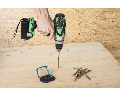 Ein Mann schraubt mit einem Akku-Bohrschrauber eine Schraube in ein Holzbrett, Schrauben und Bit-Set liegen bereit.