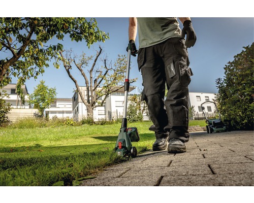 Eine Person verwendet einen Rasentrimmer im Garten, mit einem Rasenmäher im Hintergrund.