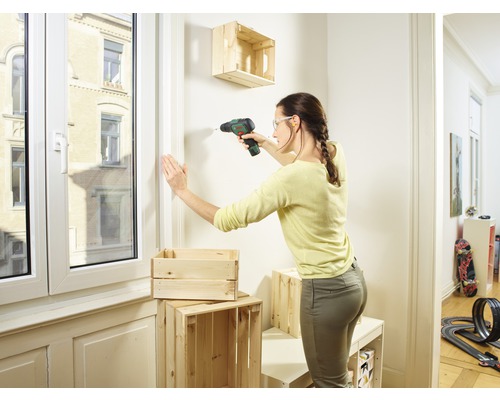 Frau bohrt Holzkiste mit Bohrmaschine an der Wand, um ein Regal zu bauen