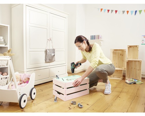 Frau arbeitet mit einem Akkubohrschrauber an einer Kiste in einem Kinderzimmer mit Puppenwagen und Schrank