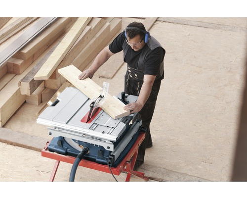 Ein Mann schneidet Holz mit einer Tischkreissäge, wobei er eine Schutzbrille und Kopfhörer trägt. Holzstapel sind im Hintergrund zu sehen.