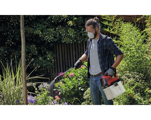 Mann mit Schutzbrille und Atemschutzmaske, der einen Akku-Pflanzensprüher im Garten verwendet.