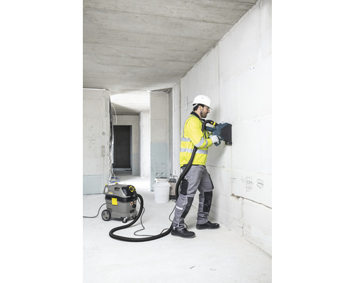 Ein Arbeiter nutzt eine Mauernutfräse an einer Wand, mit einem Staubsauger im Hintergrund auf einer Baustelle.