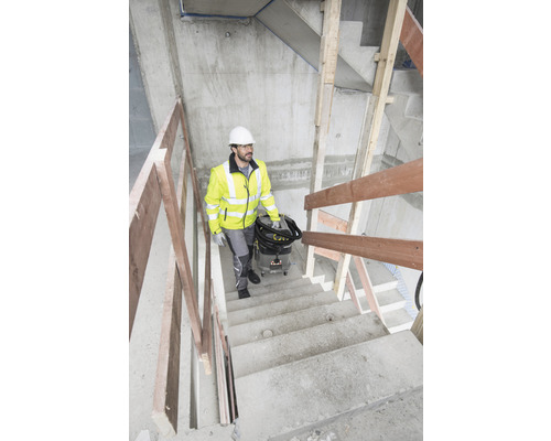 Ein Bauarbeiter mit Schutzhelm und Warnweste trägt einen Staubsauger eine Treppe hinauf.