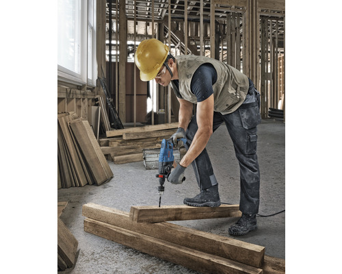 Ein Bauarbeiter bohrt mit einer Bohrmaschine ein Loch in ein Holzbrett auf einer Baustelle.