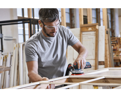 Ein Mann schleift einen Holzrahmen mit einem Exzenterschleifer in einer Werkstatt. Er trägt dabei eine Schutzbrille und Ohrstöpsel.