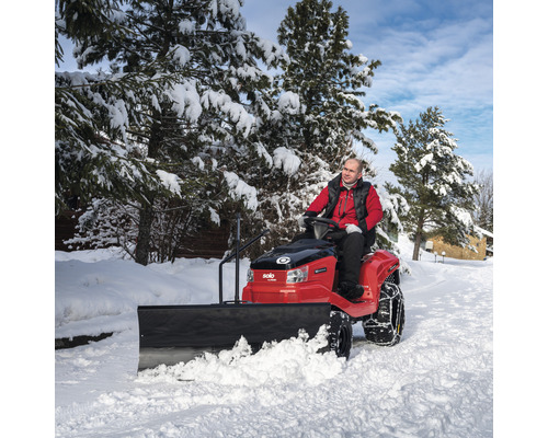 Mann fährt mit Rasentraktor und Schneeschild auf schneebedeckter Straße