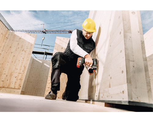 Ein Bauarbeiter mit Schutzhelm befestigt eine Holzplatte mit einem Akku-Schlagschrauber auf einer Baustelle.