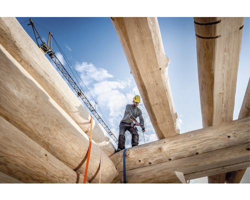Ein Mann arbeitet mit einem Akkubohrer auf einem Holzhausbau, im Hintergrund ein Baukran.