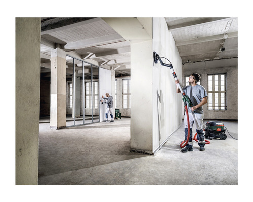 Ein Mann schleift eine Wand mit einem Langhalsschleifer in einem unrenovierten Raum. Ein Industriesauger steht daneben.