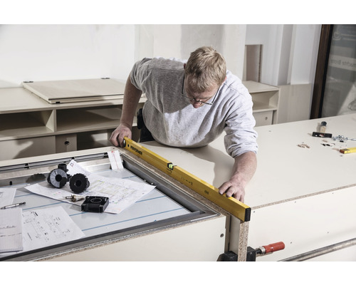 Ein Mann verwendet eine Wasserwaage auf einer Werkbank. Baupläne und Werkzeuge liegen bereit.